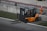 A forklift with an orange body and black details is positioned near a concrete barrier with two numbered signs. The operator inside the forklift is wearing a helmet. Nearby, a person in an orange jumpsuit and white safety helmet is standing on a paved surface. The area is bordered by grass and contains metallic railings.