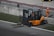 A forklift with an orange body and black details is positioned near a concrete barrier with two numbered signs. The operator inside the forklift is wearing a helmet. Nearby, a person in an orange jumpsuit and white safety helmet is standing on a paved surface. The area is bordered by grass and contains metallic railings.