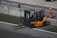 A forklift with an orange body and black details is positioned near a concrete barrier with two numbered signs. The operator inside the forklift is wearing a helmet. Nearby, a person in an orange jumpsuit and white safety helmet is standing on a paved surface. The area is bordered by grass and contains metallic railings.