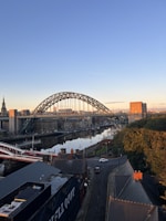 An urban landscape features a prominent arched steel bridge spanning a river, with the city skyline in the background. There are red brick buildings and a road running through a residential area in the foreground. The sky is clear with the warm glow of sunset or sunrise, casting a serene atmosphere over the scene.