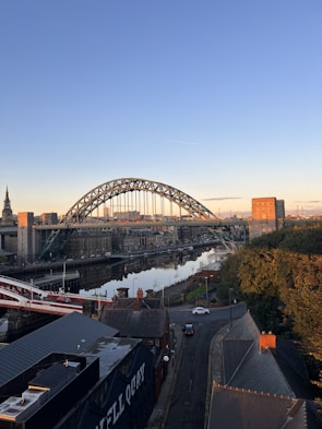An urban landscape features a prominent arched steel bridge spanning a river, with the city skyline in the background. There are red brick buildings and a road running through a residential area in the foreground. The sky is clear with the warm glow of sunset or sunrise, casting a serene atmosphere over the scene.