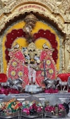 A decorated idol is positioned centrally, surrounded by vibrant colors and ornate objects. The idol is adorned with intricate jewelry and garments, set against a backdrop of rich red and yellow hues. Silver statues of cows are placed on either side, and flowers are arranged beneath the idol, adding to the decorative theme.