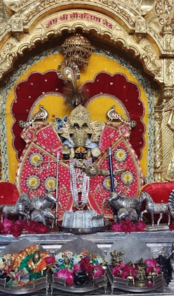 A decorated idol is positioned centrally, surrounded by vibrant colors and ornate objects. The idol is adorned with intricate jewelry and garments, set against a backdrop of rich red and yellow hues. Silver statues of cows are placed on either side, and flowers are arranged beneath the idol, adding to the decorative theme.