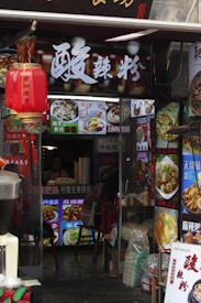 A small street food restaurant entrance with bright signage and colorful food advertisements displayed around the entrance. A red Chinese lantern hangs on the left, and several food pictures are shown. Inside, a person is visible sitting at a counter.