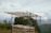 A bamboo structure stands on a hillside, surrounded by lush green vegetation and overlooking misty mountains. The sky is overcast with grey clouds.