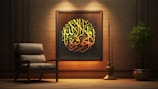 A cozy interior scene featuring a wooden wall adorned with an ornate framed piece of Arabic calligraphy. The calligraphy is illuminated with warm lighting, emphasizing its yellow and golden hues. Beside the artwork is an armchair with plush cushions, adding to the comfortable ambiance. A large, leafy plant in a pot and a tall, slender decorative element are positioned to the right, enhancing the tranquil and elegant atmosphere.