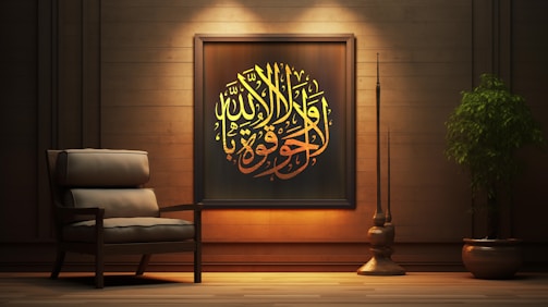 A cozy interior scene featuring a wooden wall adorned with an ornate framed piece of Arabic calligraphy. The calligraphy is illuminated with warm lighting, emphasizing its yellow and golden hues. Beside the artwork is an armchair with plush cushions, adding to the comfortable ambiance. A large, leafy plant in a pot and a tall, slender decorative element are positioned to the right, enhancing the tranquil and elegant atmosphere.