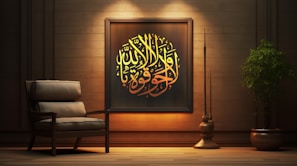A cozy interior scene featuring a wooden wall adorned with an ornate framed piece of Arabic calligraphy. The calligraphy is illuminated with warm lighting, emphasizing its yellow and golden hues. Beside the artwork is an armchair with plush cushions, adding to the comfortable ambiance. A large, leafy plant in a pot and a tall, slender decorative element are positioned to the right, enhancing the tranquil and elegant atmosphere.