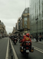 A procession of motorcyclists in uniform rides down a city street lined with historic buildings and modern glass structures. The riders wear red jackets with inscriptions, suggesting they are part of an organized group. Pedestrians watch from the sidewalks.