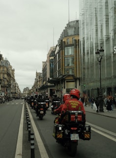 A procession of motorcyclists in uniform rides down a city street lined with historic buildings and modern glass structures. The riders wear red jackets with inscriptions, suggesting they are part of an organized group. Pedestrians watch from the sidewalks.