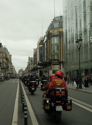 A procession of motorcyclists in uniform rides down a city street lined with historic buildings and modern glass structures. The riders wear red jackets with inscriptions, suggesting they are part of an organized group. Pedestrians watch from the sidewalks.