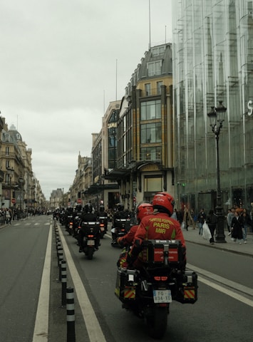 A procession of motorcyclists in uniform rides down a city street lined with historic buildings and modern glass structures. The riders wear red jackets with inscriptions, suggesting they are part of an organized group. Pedestrians watch from the sidewalks.