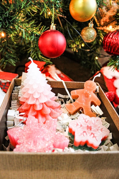 A decorative box filled with festive candles, shaped like a pink Christmas tree, a gingerbread man, and a snowflake rests beneath a Christmas tree adorned with red and gold ornaments. The box is lined with shredded paper.
