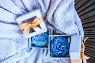 Gift boxes with blue rose-shaped candles are placed on a textured, fluffy blanket. Each box is adorned with a gold ribbon and contains a carefully crafted candle resembling a rose.