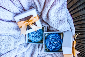 Gift boxes with blue rose-shaped candles are placed on a textured, fluffy blanket. Each box is adorned with a gold ribbon and contains a carefully crafted candle resembling a rose.