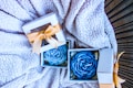 Gift boxes with blue rose-shaped candles are placed on a textured, fluffy blanket. Each box is adorned with a gold ribbon and contains a carefully crafted candle resembling a rose.