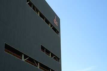 A modern building facade featuring vertical lines and rectangular windows. The structure has a dark exterior with a symbol or logo near the top. The sky is clear and bright, occupying a significant portion of the image.