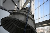 A large industrial metal structure, possibly part of a rocket engine or machinery, is displayed indoors. The structure is positioned near expansive glass windows that allow natural light to illuminate the area. The lighting casts detailed shadows on the metal surface, highlighting its grooves and texture.