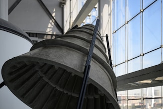 A large industrial metal structure, possibly part of a rocket engine or machinery, is displayed indoors. The structure is positioned near expansive glass windows that allow natural light to illuminate the area. The lighting casts detailed shadows on the metal surface, highlighting its grooves and texture.