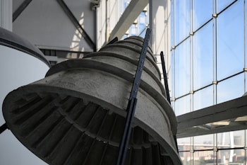 A large industrial metal structure, possibly part of a rocket engine or machinery, is displayed indoors. The structure is positioned near expansive glass windows that allow natural light to illuminate the area. The lighting casts detailed shadows on the metal surface, highlighting its grooves and texture.