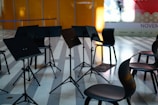 Several music stands and chairs are arranged in a semi-circle on a striped floor, suggesting a setting for a rehearsal or performance. The background includes an orange wall and a partially visible billboard with the word 'NOVEMBER' and other text.