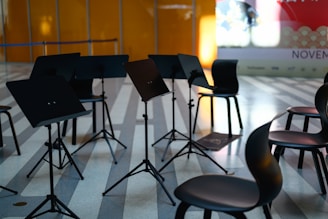 Several music stands and chairs are arranged in a semi-circle on a striped floor, suggesting a setting for a rehearsal or performance. The background includes an orange wall and a partially visible billboard with the word 'NOVEMBER' and other text.