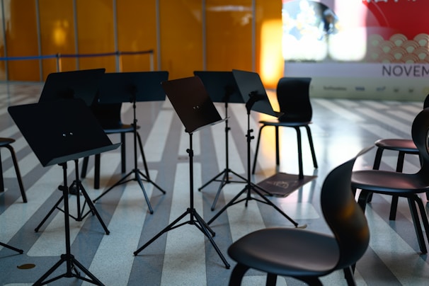 Several music stands and chairs are arranged in a semi-circle on a striped floor, suggesting a setting for a rehearsal or performance. The background includes an orange wall and a partially visible billboard with the word 'NOVEMBER' and other text.