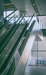 A modern indoor space features a pair of parallel escalators leading up to an upper floor. The design includes glass panels and sleek metal railings. Large glass windows in the background provide ample natural light, creating a bright and airy atmosphere.