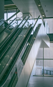 A modern indoor space features a pair of parallel escalators leading up to an upper floor. The design includes glass panels and sleek metal railings. Large glass windows in the background provide ample natural light, creating a bright and airy atmosphere.