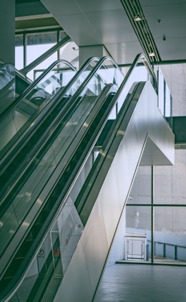 A modern indoor space features a pair of parallel escalators leading up to an upper floor. The design includes glass panels and sleek metal railings. Large glass windows in the background provide ample natural light, creating a bright and airy atmosphere.