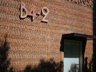 A textured brick wall with an intriguing pattern, featuring a metal sign displaying 'B4-2' mounted on it. A small overhang or canopy shades a glass door below, casting a shadow on the surface. Shadows of trees or plants are also visible on the wall, adding to the dynamics of the scene.