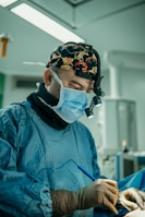 A surgeon wearing a surgical mask, cap with a pattern, and operating garments is focused intently on a procedure. Surgical lights are illuminating the scene, and various medical equipment is visible in the background.