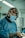 A surgeon wearing a surgical mask, cap with a pattern, and operating garments is focused intently on a procedure. Surgical lights are illuminating the scene, and various medical equipment is visible in the background.