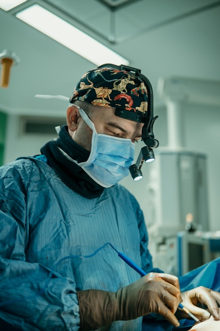 A surgeon wearing a surgical mask, cap with a pattern, and operating garments is focused intently on a procedure. Surgical lights are illuminating the scene, and various medical equipment is visible in the background.