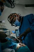 A surgeon wearing a mask and headlamp is focused on performing a procedure in an operating room. The room is dimly lit, and the patient is lying on an operating table, covered with a sterile cloth. Instruments and medical equipment are visible in the background.
