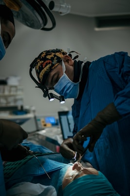 A surgeon wearing a mask and headlamp is focused on performing a procedure in an operating room. The room is dimly lit, and the patient is lying on an operating table, covered with a sterile cloth. Instruments and medical equipment are visible in the background.