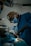 A surgeon wearing a mask and headlamp is focused on performing a procedure in an operating room. The room is dimly lit, and the patient is lying on an operating table, covered with a sterile cloth. Instruments and medical equipment are visible in the background.