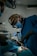 A surgeon wearing a mask and headlamp is focused on performing a procedure in an operating room. The room is dimly lit, and the patient is lying on an operating table, covered with a sterile cloth. Instruments and medical equipment are visible in the background.