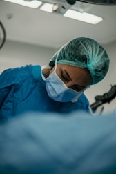 A medical professional is wearing a blue surgical gown, a hair net, and a face mask, concentrating on a task in a hospital or operating room setting.