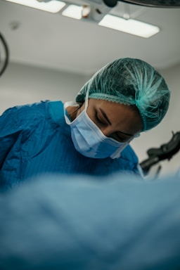 A medical professional is wearing a blue surgical gown, a hair net, and a face mask, concentrating on a task in a hospital or operating room setting.