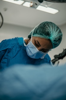 A medical professional is wearing a blue surgical gown, a hair net, and a face mask, concentrating on a task in a hospital or operating room setting.