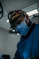 A person wearing surgical attire and a face mask, utilizing a magnifying headlamp in a medical setting. The environment appears sterile with dim lighting, highlighting the focus on precision and skill. The individual is deeply concentrated on their task.