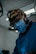 A person wearing surgical attire and a face mask, utilizing a magnifying headlamp in a medical setting. The environment appears sterile with dim lighting, highlighting the focus on precision and skill. The individual is deeply concentrated on their task.