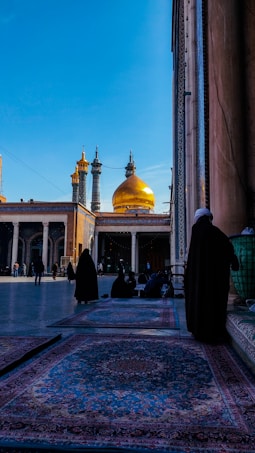 The image depicts a courtyard of a mosque featuring a vibrant blue sky above. The focal point of the image is the golden dome of the mosque, flanked by two smaller, intricately designed towers. Within the courtyard, several people dressed in traditional attire are either walking or sitting on ornate rugs that cover part of the ground.