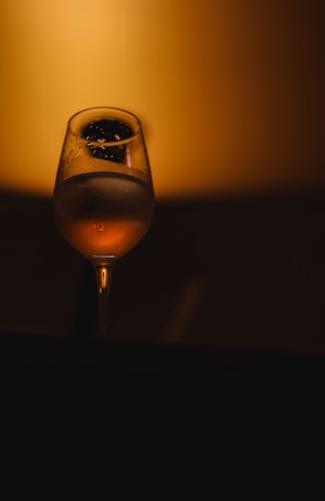 A wine glass containing liquid with condensation droplets, set against a warm, softly illuminated amber background. The glass reflects subtle highlights, creating an elegant and minimalistic impression.