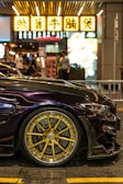 A sleek, dark-colored sports car is parked on a street at night. The focus is on its gold, intricately designed alloy wheels. In the background, there are illuminated signs with text in an Asian script, typical of a bustling urban environment. Several people can be seen walking, adding a sense of liveliness to the scene.