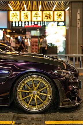 A sleek, dark-colored sports car is parked on a street at night. The focus is on its gold, intricately designed alloy wheels. In the background, there are illuminated signs with text in an Asian script, typical of a bustling urban environment. Several people can be seen walking, adding a sense of liveliness to the scene.