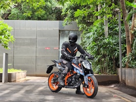 A person wearing black motorcycle gear, including a helmet and gloves, is seated on a brightly colored sports motorcycle with orange wheels. The setting is an outdoor area with lush green foliage, a concrete wall, and gray tiled pavement.