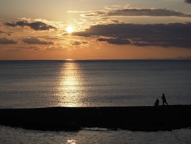 The image depicts a serene sunset over a calm ocean with shades of orange and gold reflected on the water. Silhouettes of two people are visible on the darkened shore in the lower right corner.