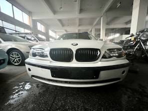 A white car is parked indoors in what appears to be a garage or showroom. The setting includes other parked cars and a motorcycle visible in the background. The environment is well-lit with large windows, and the floor shows some reflections from water or oil. The car's front view is prominently displayed with emphasis on its grille and headlights.